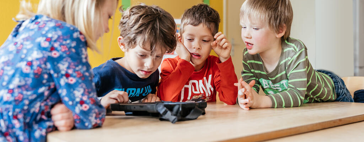 Kinder sitzen gemeinsam an einem Tisch und entdecken Inhalte auf einem Tablet.